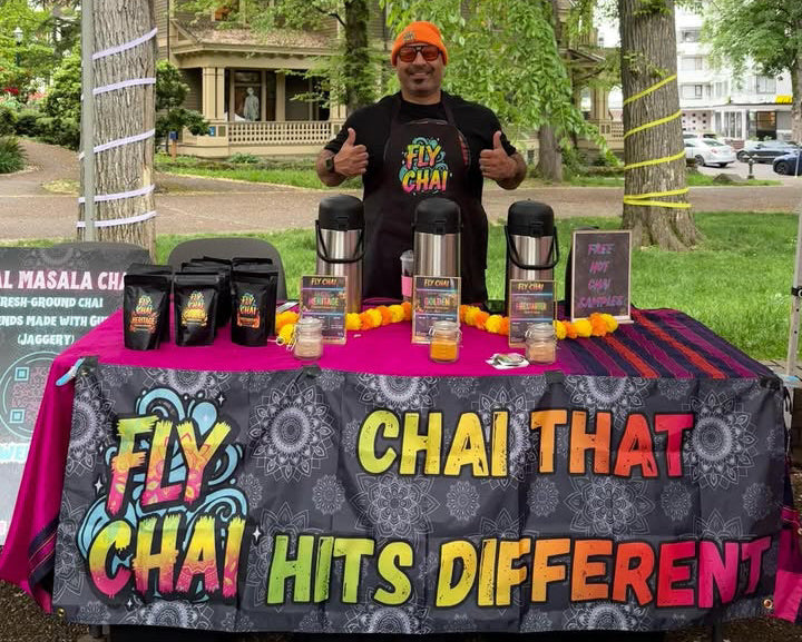 A smiling vendor giving a double thumbs-up behind a vibrant Fly Chai market stall. The table is dressed in a hot pink tablecloth and a black banner that reads "FLY CHAI — CHAI THAT HITS DIFFERENT" in bold, colorful lettering. On the table are three large stainless steel beverage dispensers labeled Heritage, Golden, and FireStarter, alongside packaged loose-leaf chai blends and a sign offering "Free Hot Chai Samples.