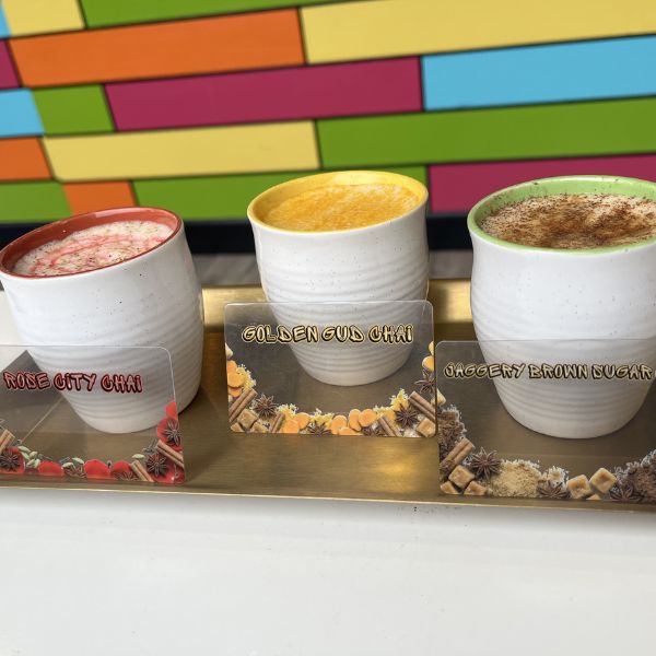 Three ceramic cups filled with different flavored coffee drinks arranged on a tray in front of a colorful geometric wall. The cups are labeled Rose City Chai, Golden Cua Chai, and Jaccerly Brimmm Sugar.