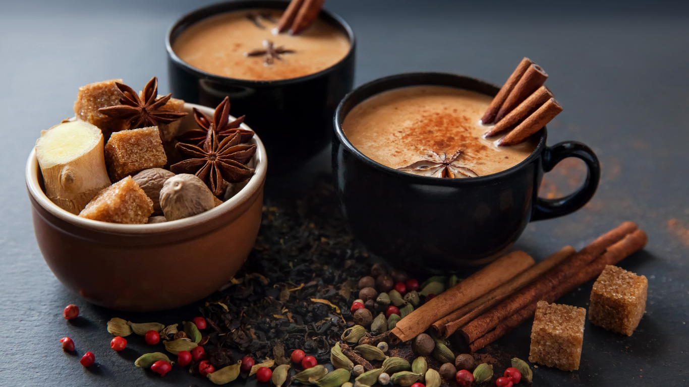 Two black mugs of hot chocolate with cinnamon sticks, a bowl of spices, and a dark background.