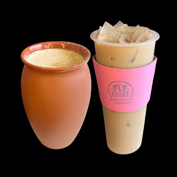 A terracotta clay cup filled with a light brown beverage sits beside a clear plastic iced coffee cup with a pink Fly Chai sleeve, both photographed against a solid black background.