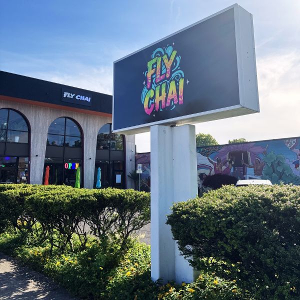 A large outdoor digital billboard mounted on a white post, displaying the colorful 'FLY CHAI' logo. The sign is situated in a landscaped garden area with green hedges, with a modern building and a mural visible in the background under a blue sky.