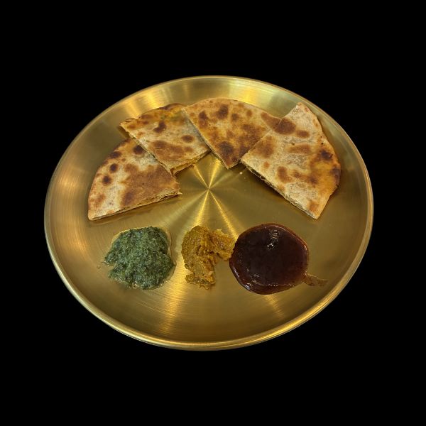 A round brushed gold metal plate containing three triangular pieces of toasted flatbread, a dollop of green chutney, a dollop of yellow curry powder, and a dollop of dark red chutney, set against a solid black background.