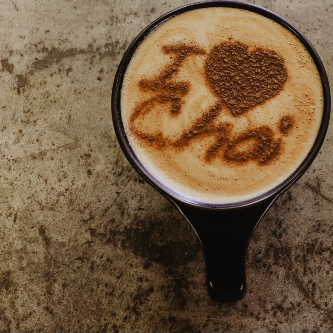 A black ceramic mug filled with a cappuccino or latte, viewed from above. The foam on top has latte art spelling 'I <heart> Chai' in brown cocoa powder, set against a textured concrete-like surface.