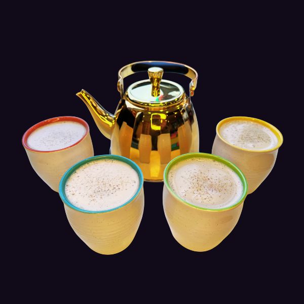 A polished gold teapot with a matching lid, surrounded by four small ceramic cups with colorful rims (red, green, yellow, and teal) filled with a frothy beverage, set against a solid black background.
