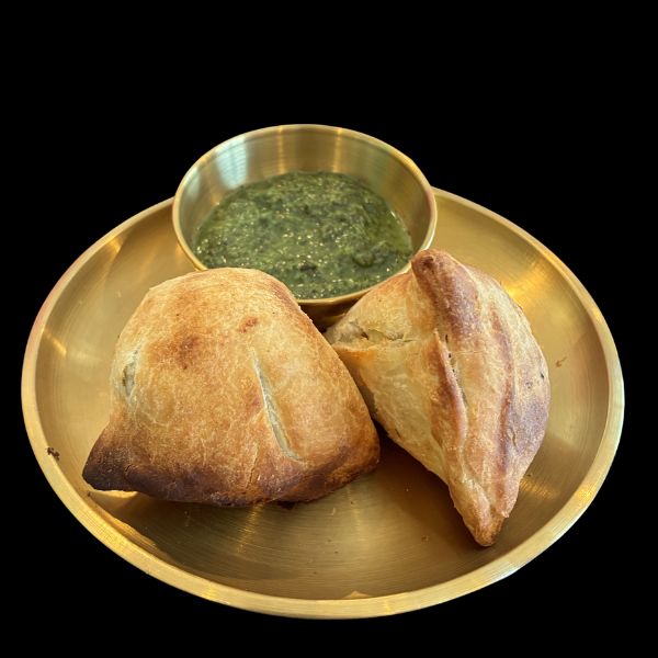 Two golden-brown baked pastries on a round brass plate with a small brass bowl of green chutney, set against a solid black background.